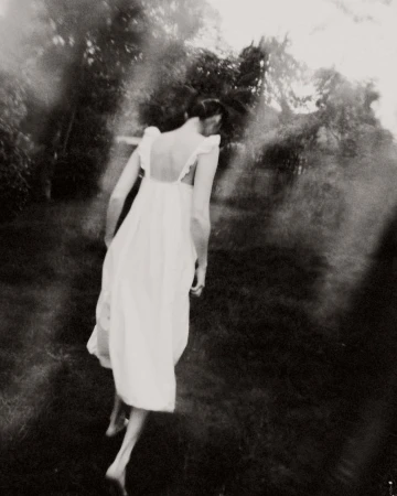 Woman in a long white dress walking through a dark garden captured in blurry monochromatic style