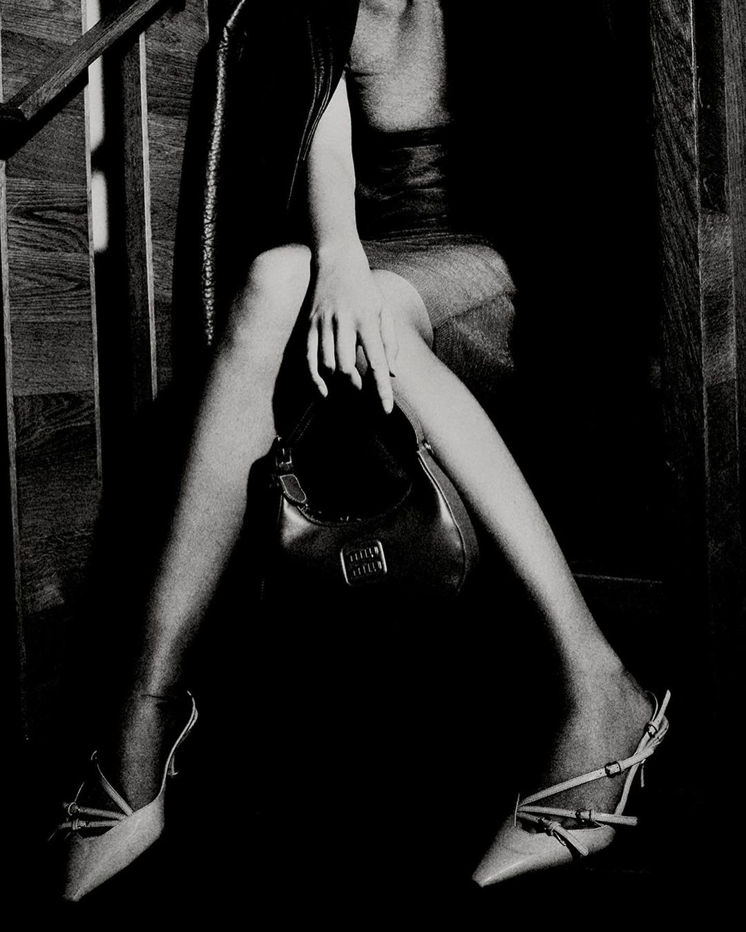A woman is sitting on wooden steps, modeling high-heeled shoes and holding a purse with the 'MIU MIU' logo. The image features a dark, moody, black-and-white aesthetic.