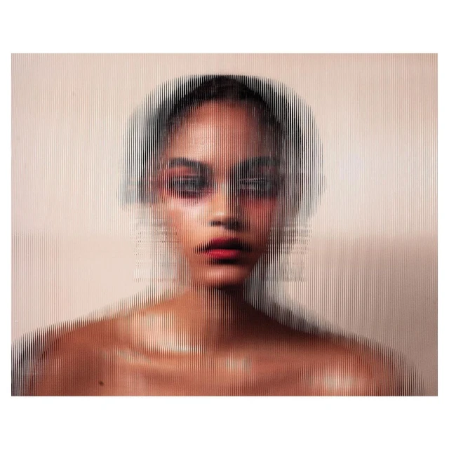 Portrait of a woman with red eyeshadow and lipstick, partially obscured by a clear, vertically-grooved material creating a diffused effect.
