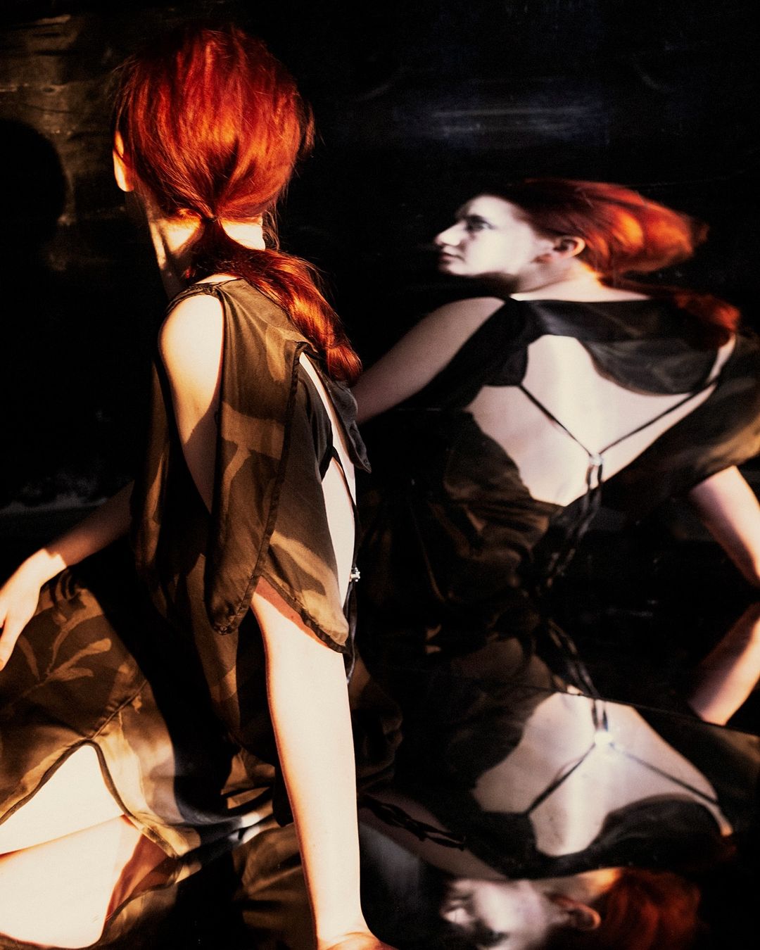 Two red-haired women in sheer black dresses pose in a dimly lit setting, one facing away and the other reflected, creating a striking image.
