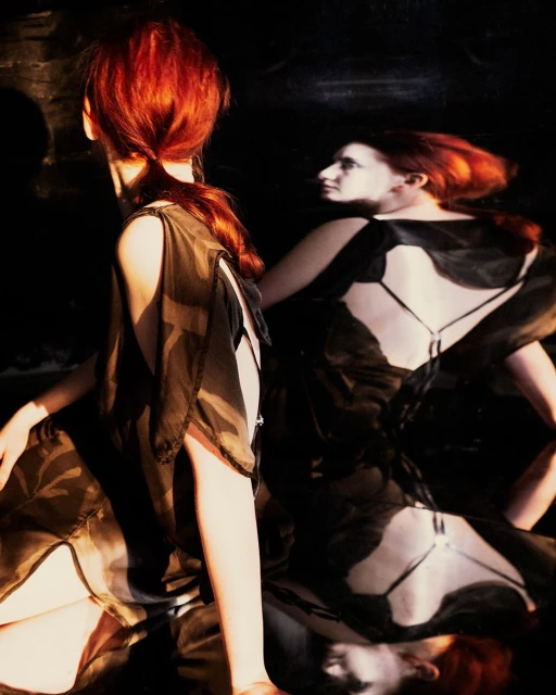 Two red-haired women in sheer black dresses pose in a dimly lit setting, one facing away and the other reflected, creating a striking image.