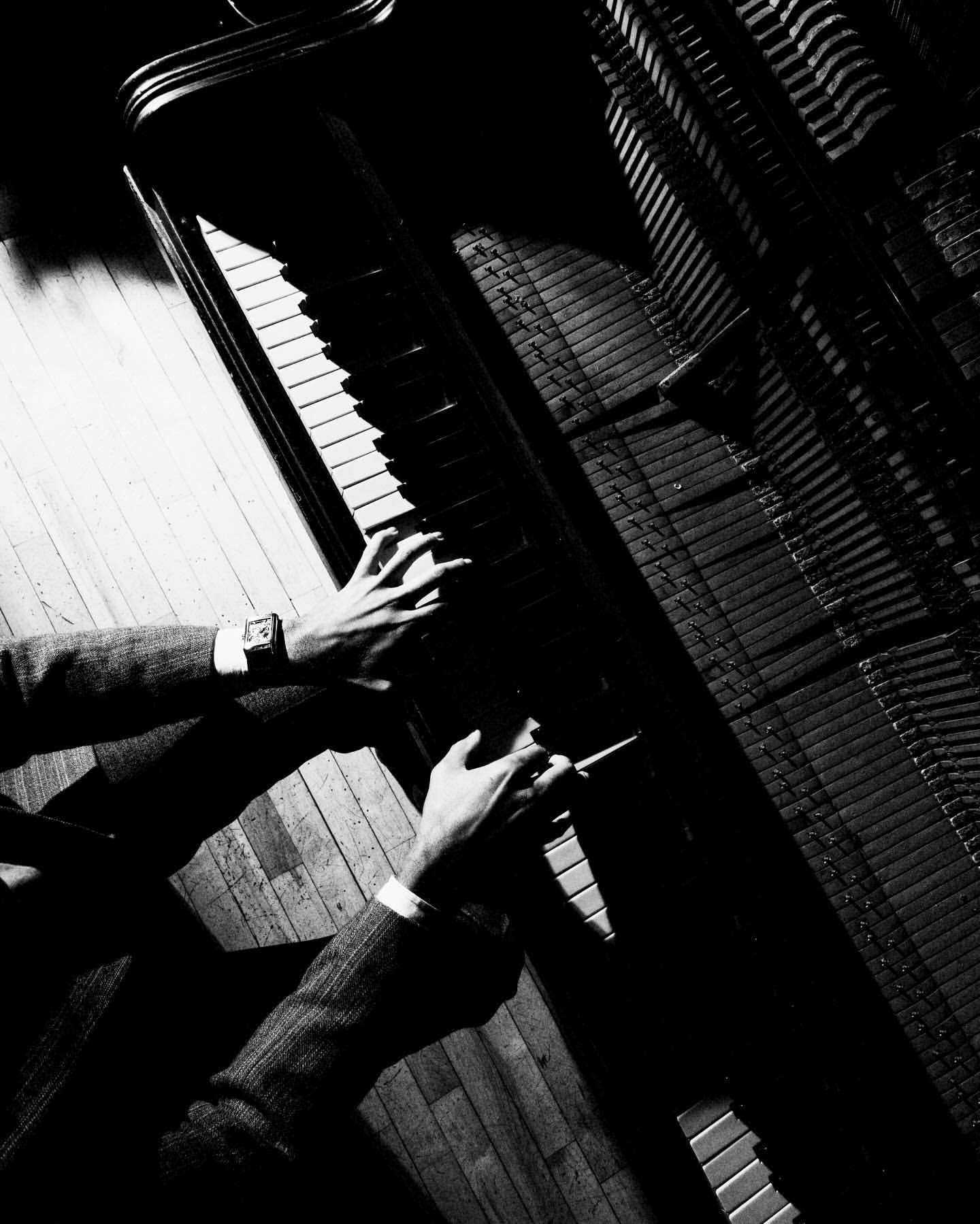 A pair of hands play an upright piano in a high-contrast black-and-white photograph, showing a suit jacket and watch in a wood-floored room.