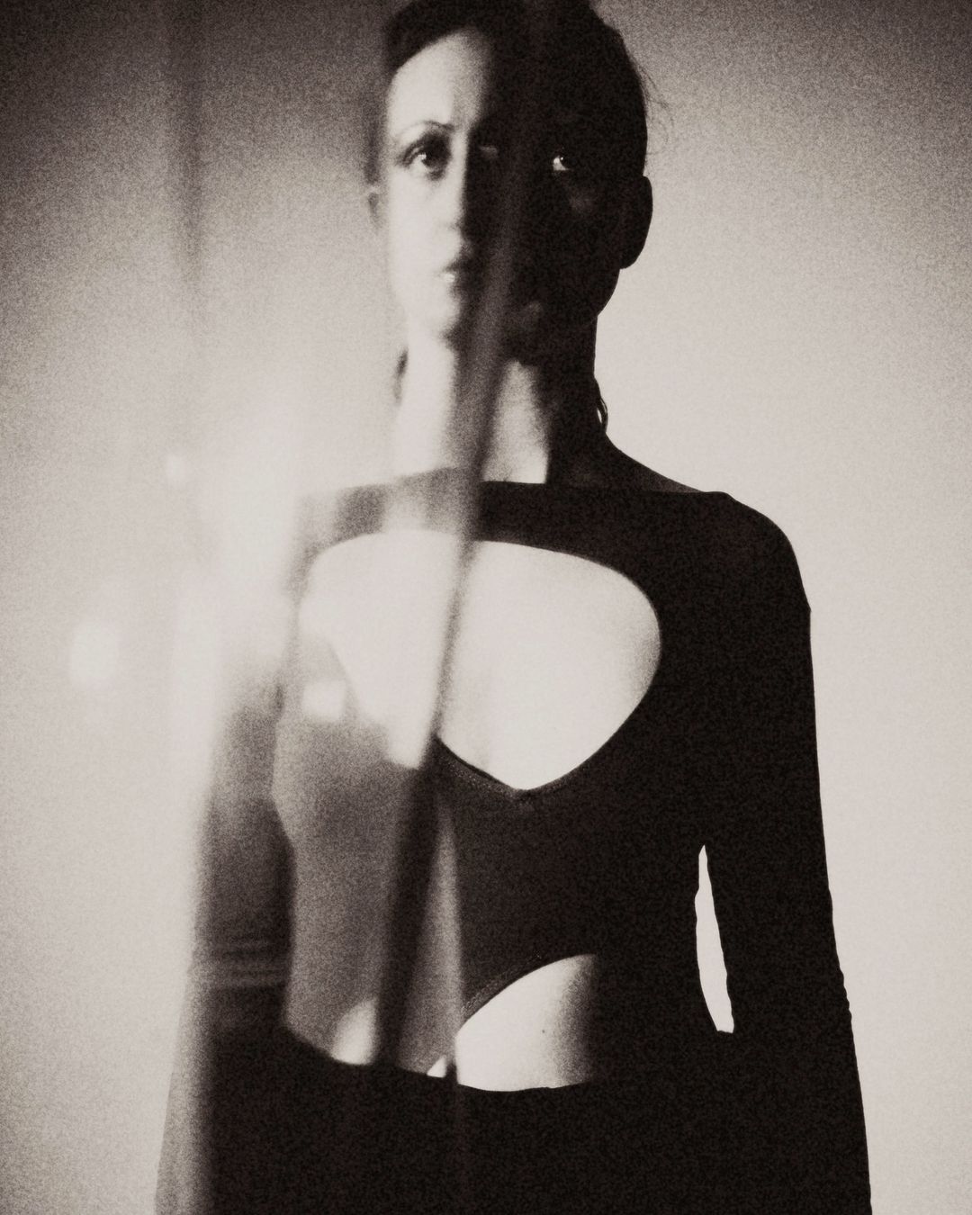 Monochrome portrait of a slender adult woman with a neutral expression wearing a long-sleeved top with chest cutouts, indoors.