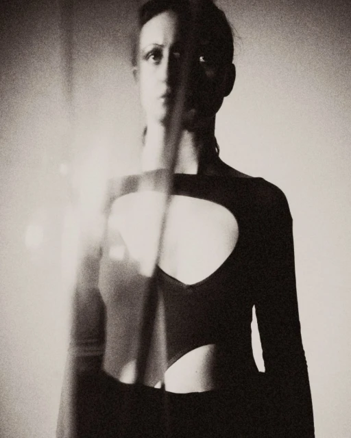 Monochrome portrait of a slender adult woman with a neutral expression wearing a long-sleeved top with chest cutouts, indoors.