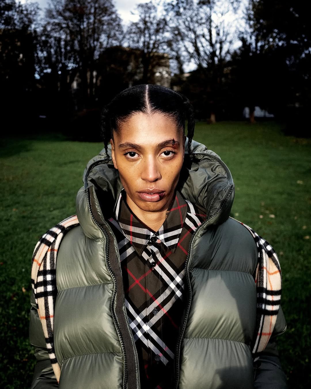 Close-up portrait of a person wearing a quilted puffer vest and plaid shirt, standing outdoors with a blurred grassy and treed background.