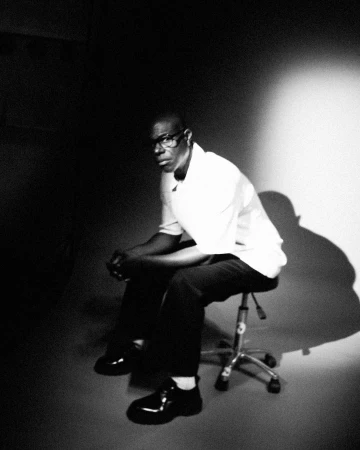 Black man wearing glasses and a light short-sleeve shirt seated on a rolling studio stool against a light wall