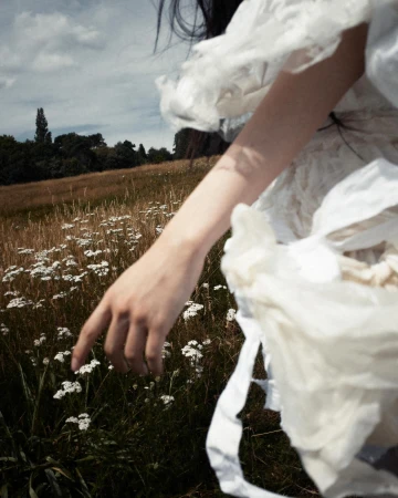 Close-up of a person reaching out over a field of wild white flowers wearing a textured white garment