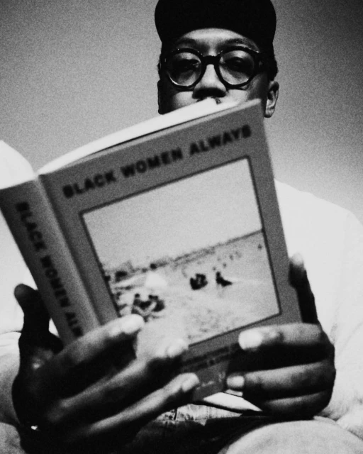 Black and white portrait of a man reading a book titled 'Black Women Always,' wearing glasses and a baseball cap, close-up.