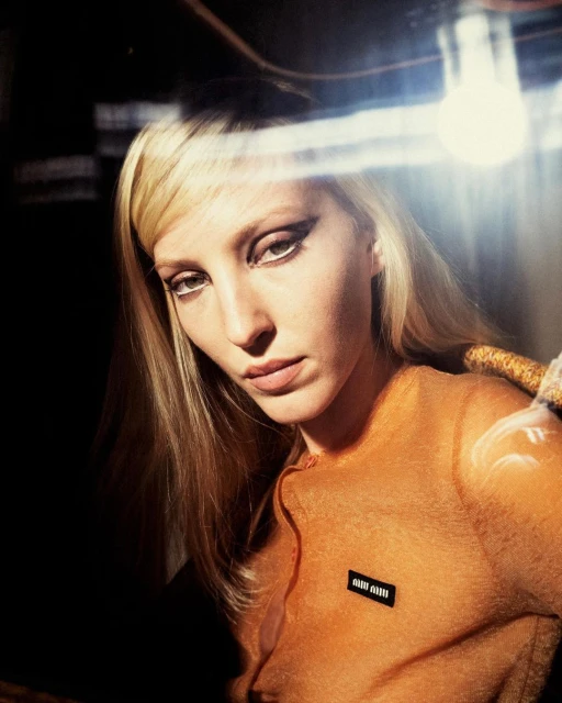 Close-up portrait of a blonde woman with striking eye makeup wearing an orange sweater, with light flares on one side of the frame.