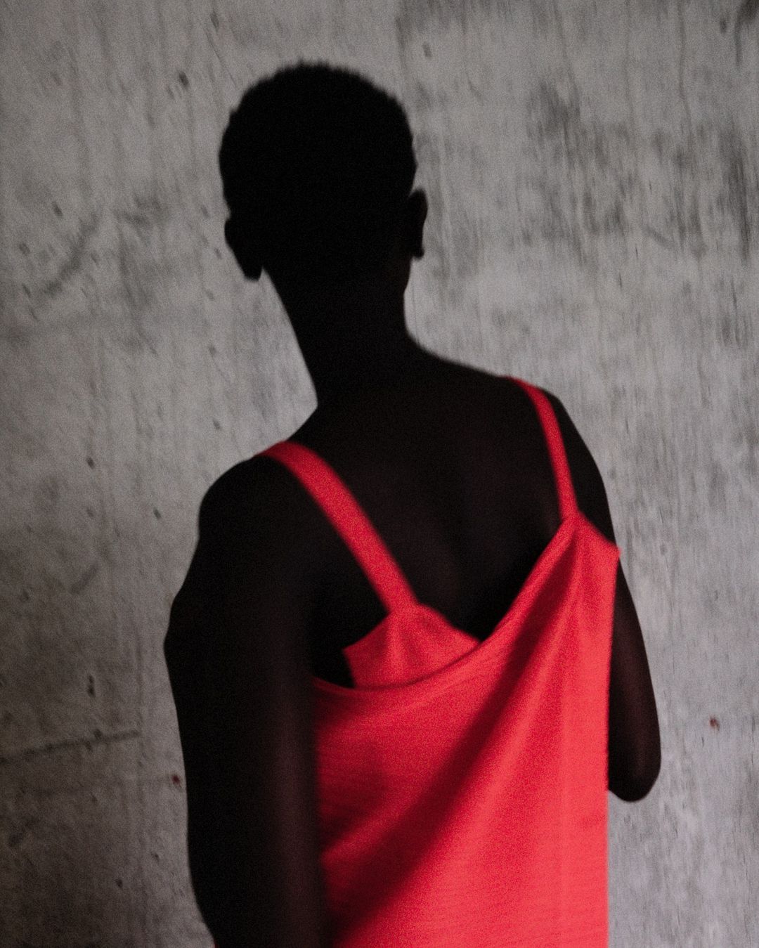 Silhouette of a person with short hair wearing a red sleeveless top against a textured concrete wall.
