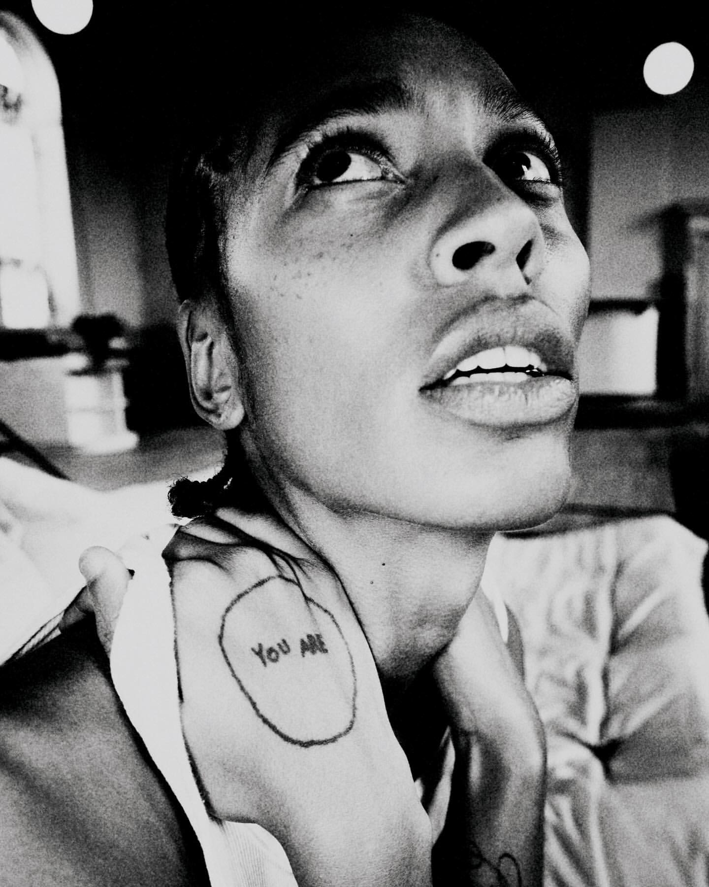 Monochromatic portrait of an adult woman with short hair looking upwards, featuring a visible tattoo on her hand and upper body.