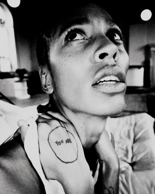 Monochromatic portrait of an adult woman with short hair looking upwards, featuring a visible tattoo on her hand and upper body.