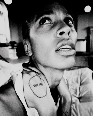 Monochromatic portrait of an adult woman with short hair looking upwards, featuring a visible tattoo on her hand and upper body.