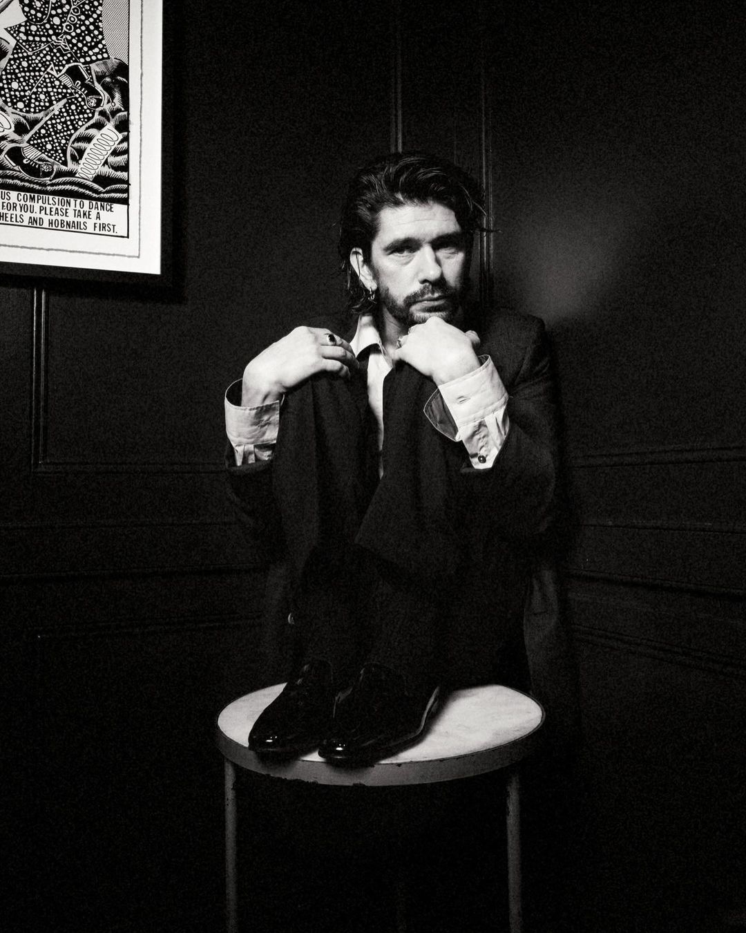 Black and white portrait of an adult male in a suit, sitting on a stool, in front of a dark textured wall with framed art.