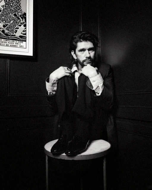 Black and white portrait of an adult male in a suit, sitting on a stool, in front of a dark textured wall with framed art.