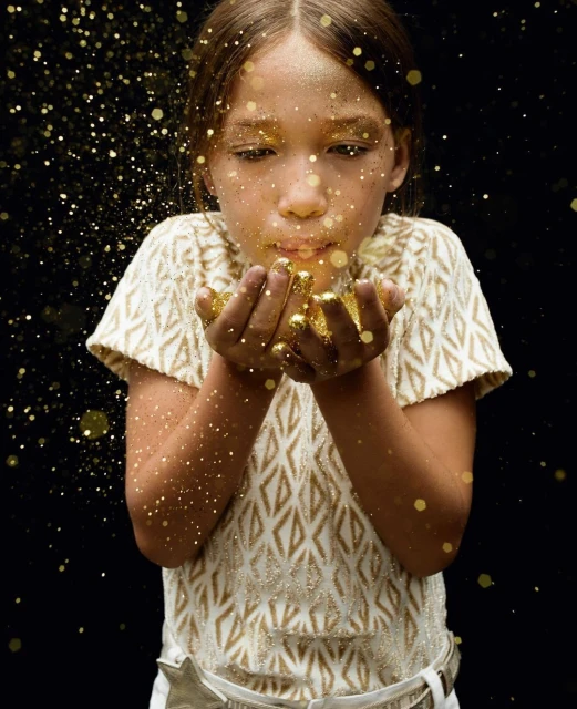 Young girl blows gold glitter toward the camera in a studio shot with a dark background, creating a magical, sparkling effect.
