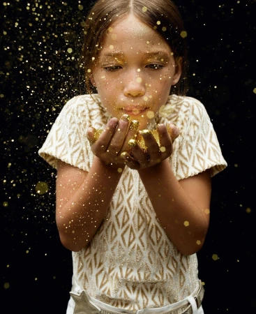 Young girl blows gold glitter toward the camera in a studio shot with a dark background, creating a magical, sparkling effect.