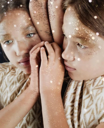 Close-up portrait of a young person with gold glitter on face and hands, reflected in a mirror with bokeh lights.