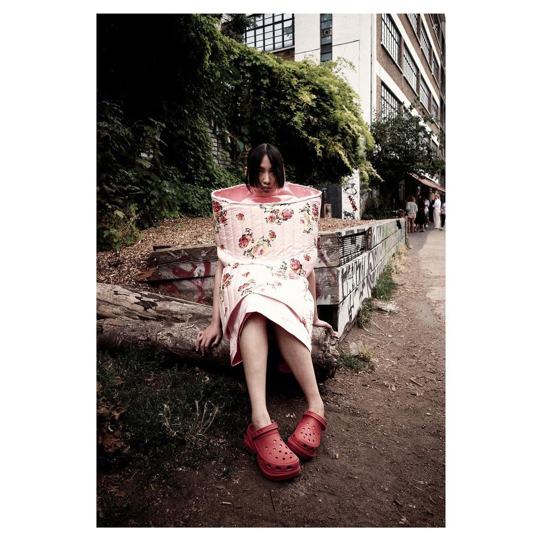 A woman poses in an unusual floral-print outfit, sitting on a log in a park-like setting near an urban building on a sunny day.