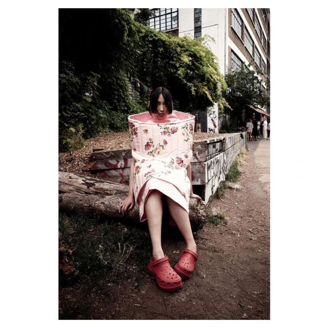 A woman poses in an unusual floral-print outfit, sitting on a log in a park-like setting near an urban building on a sunny day.