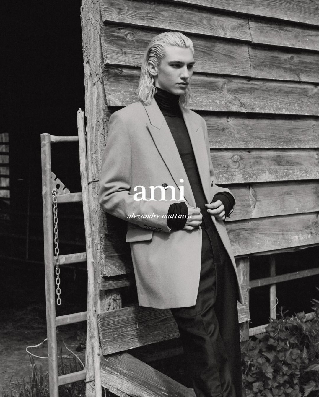 A blonde male fashion model poses in front of a weathered wood wall, wearing a black turtleneck and tan blazer in a black and white portrait.