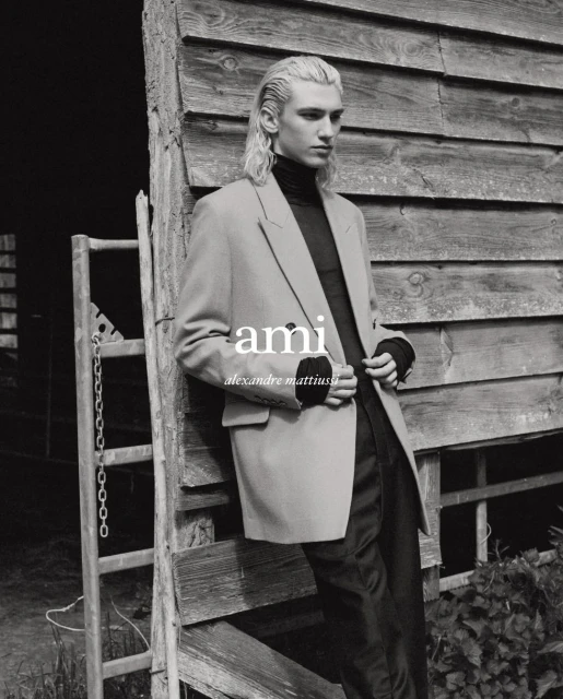 A blonde male fashion model poses in front of a weathered wood wall, wearing a black turtleneck and tan blazer in a black and white portrait.