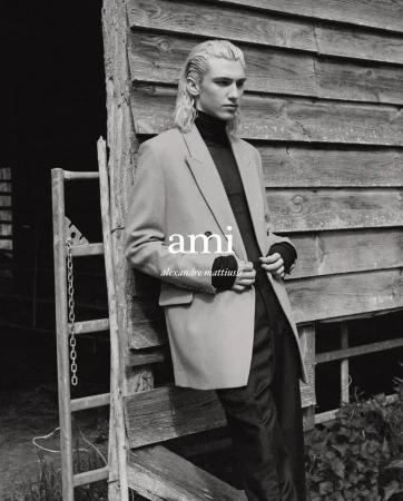 A blonde male fashion model poses in front of a weathered wood wall, wearing a black turtleneck and tan blazer in a black and white portrait.
