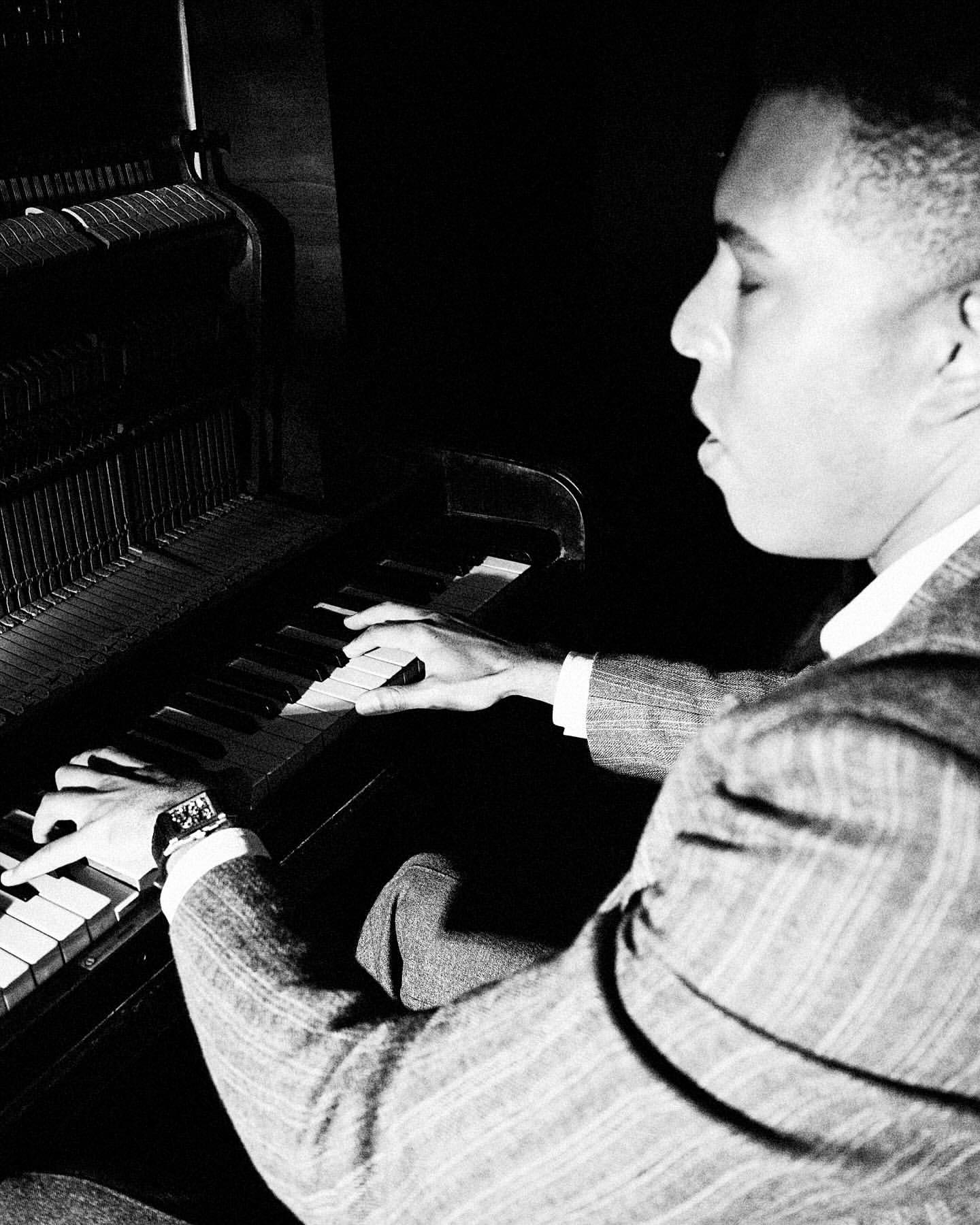 African American man plays the piano in a dimly lit room, wearing a suit with a watch on his left wrist.