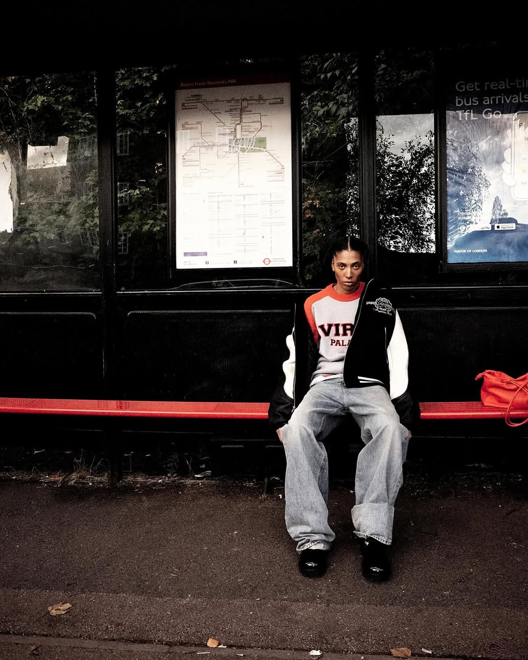 Young adult sits at bus stop in streetwear style. Streetwear fashion: varsity jacket, wide-leg denim jeans, platform shoes, bus route map.