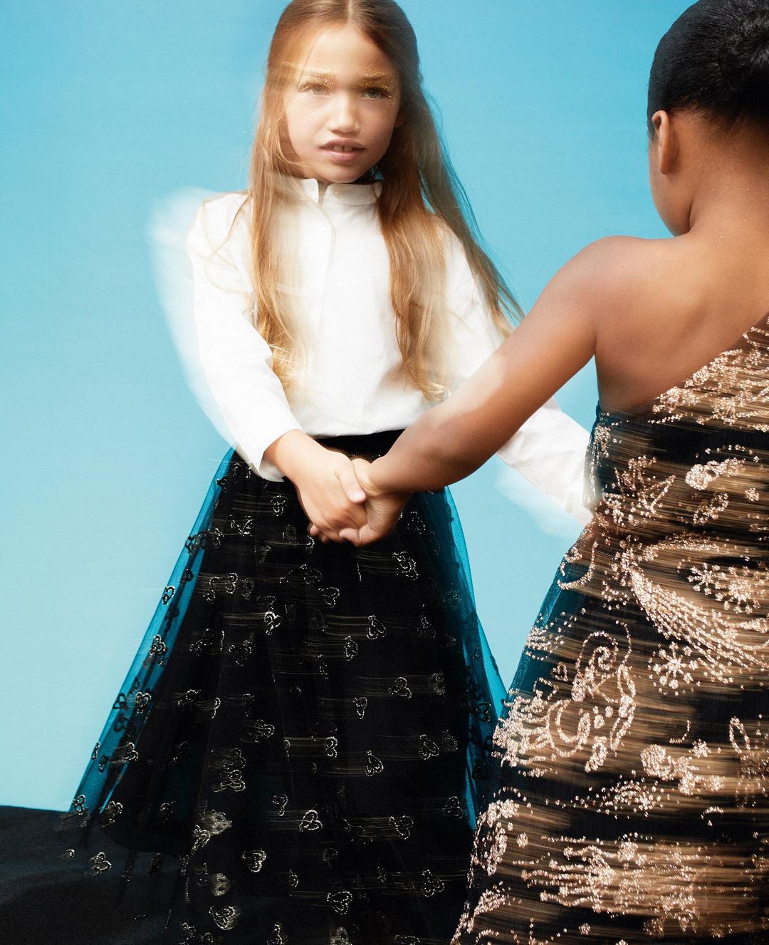 Two young girls in formal gowns hold hands against a light blue backdrop, one in white and black, the other in gold and black.