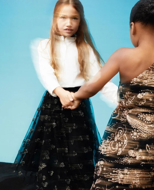 Two young girls in formal gowns hold hands against a light blue backdrop, one in white and black, the other in gold and black.