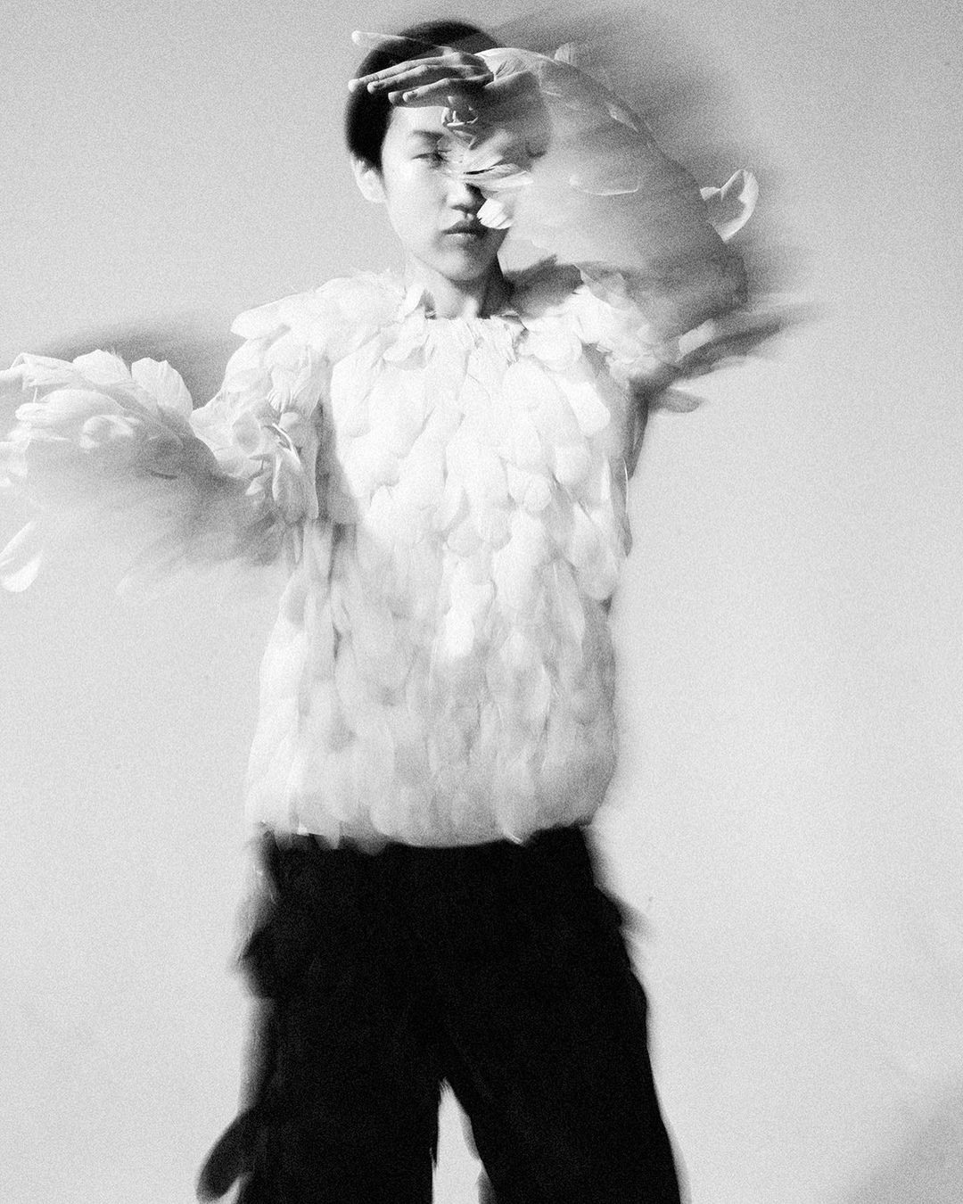 Monochromatic image of an Asian woman in a feathered garment, standing indoors with hands raised, showcasing unique fashion and artistic expression.