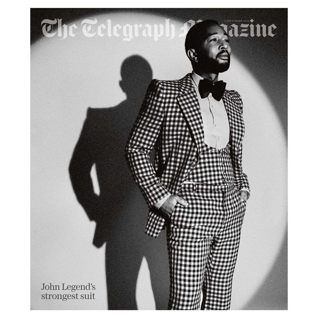 John Legend in a black and white magazine cover photo. He is wearing a checkered suit, vest, bow tie, and a white dress shirt.