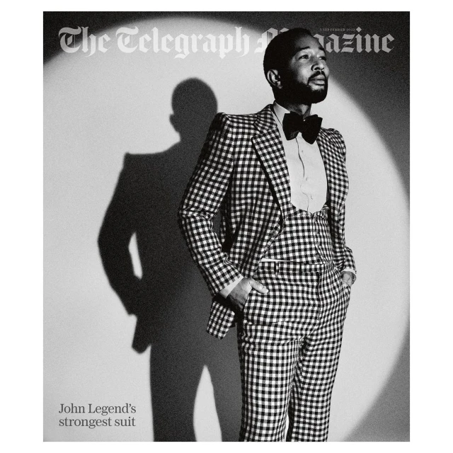 John Legend in a black and white magazine cover photo. He is wearing a checkered suit, vest, bow tie, and a white dress shirt.
