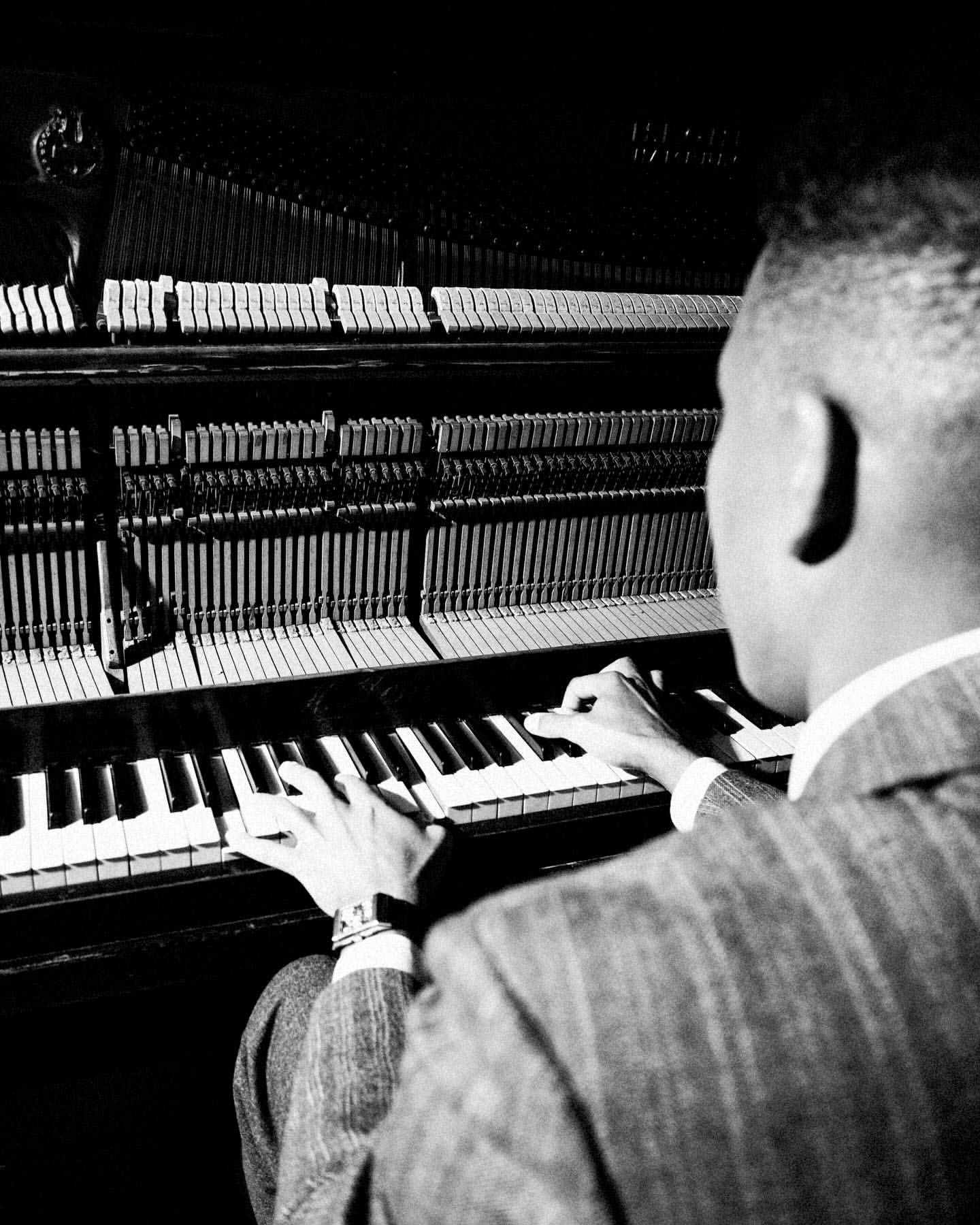 Monochrome portrait of an adult playing an open upright piano. Man wears suit jacket. The image features a close camera angle.