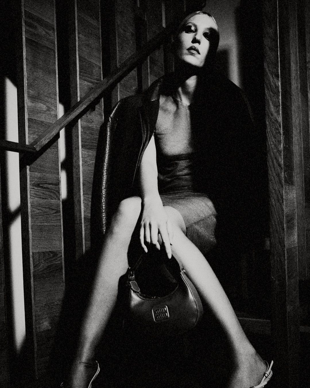 Monochrome fashion shot of a woman seated on wooden stairs, wearing a jacket and holding a Miu Miu bag, with a staircase background.