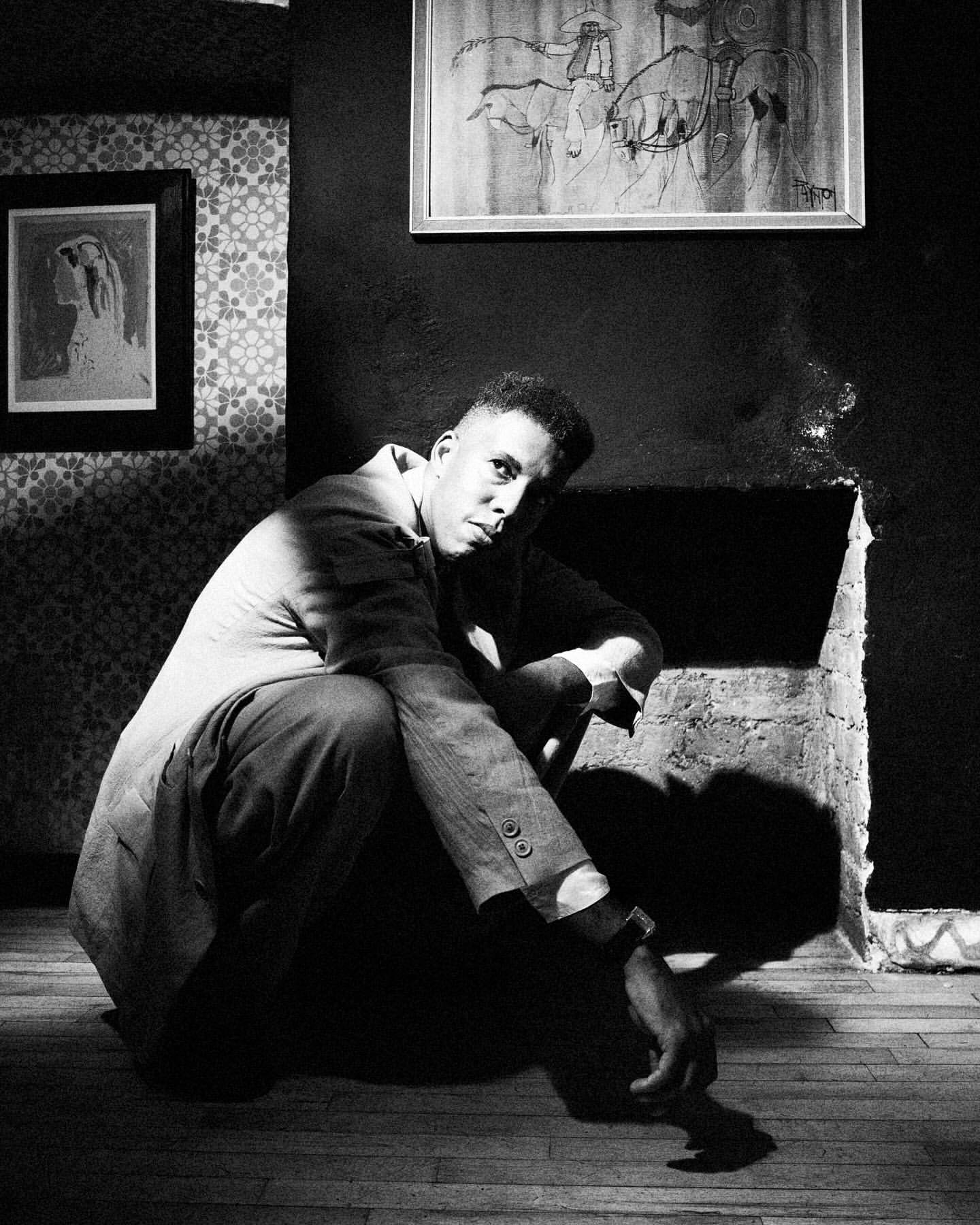 Monochrome portrait of a man crouching in front of a fireplace with artwork in the background. The image is in black and white.