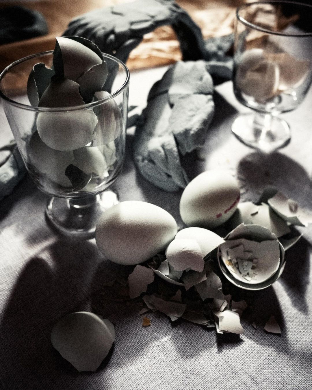 Close-up of white chicken eggs, some cracked, in a clear stemmed glass and on a linen surface with shadows, in natural lighting.