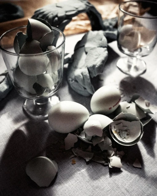 Close-up of white chicken eggs, some cracked, in a clear stemmed glass and on a linen surface with shadows, in natural lighting.