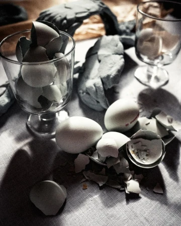 Close-up of white chicken eggs, some cracked, in a clear stemmed glass and on a linen surface with shadows, in natural lighting.