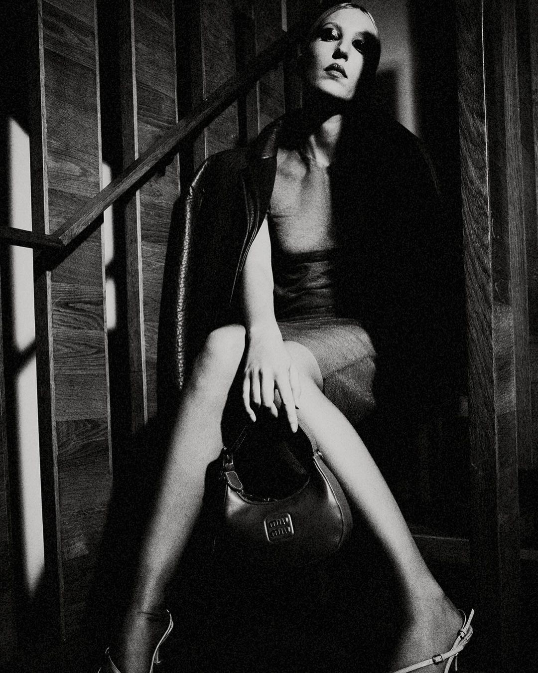 A striking black and white fashion shot of a stylish woman with a handbag, sitting on wooden stairs, wearing a dress and jacket.