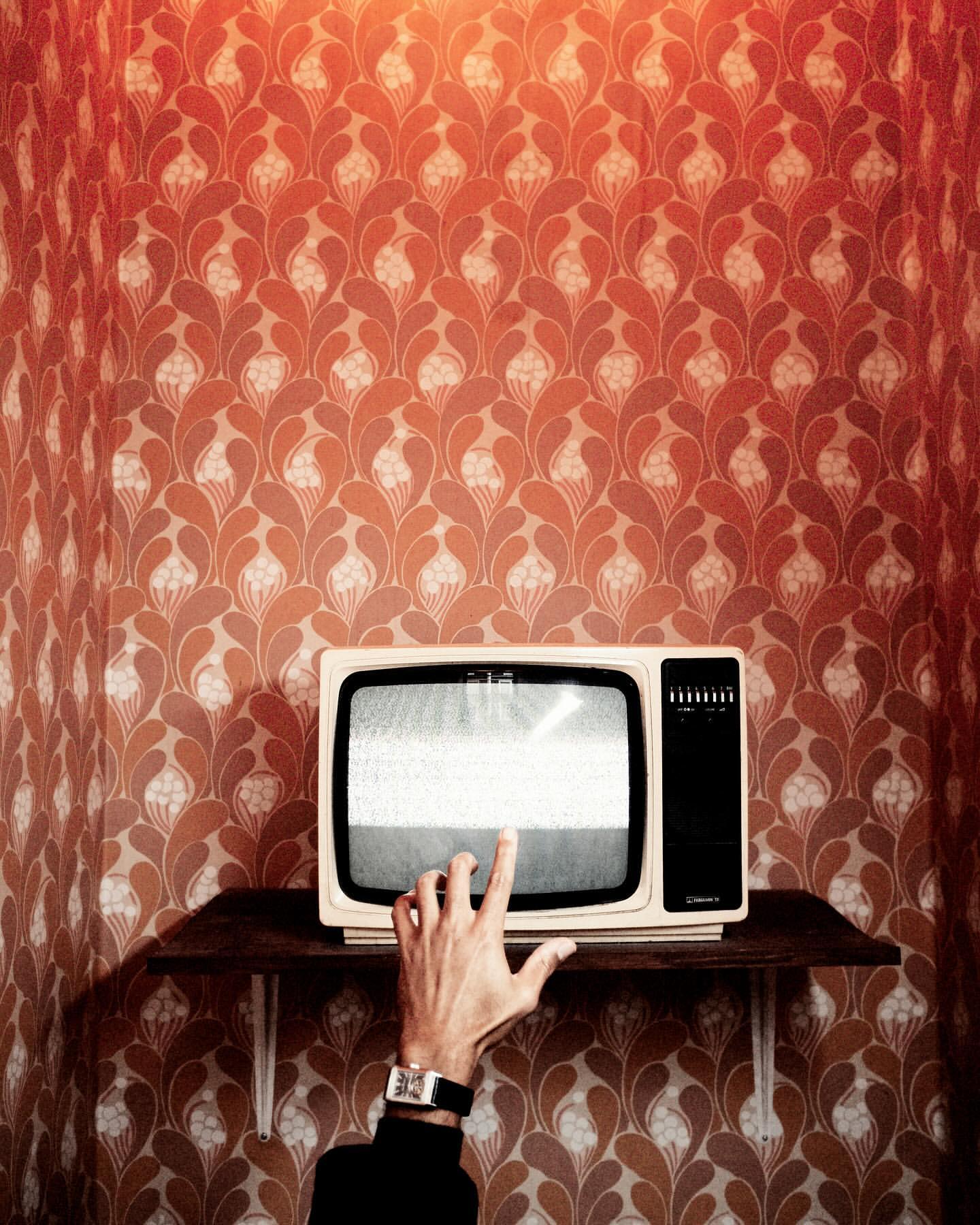 Vintage TV on a shelf with an arm reaching to turn it on in a room with patterned wallpaper, creating a retro-themed aesthetic.