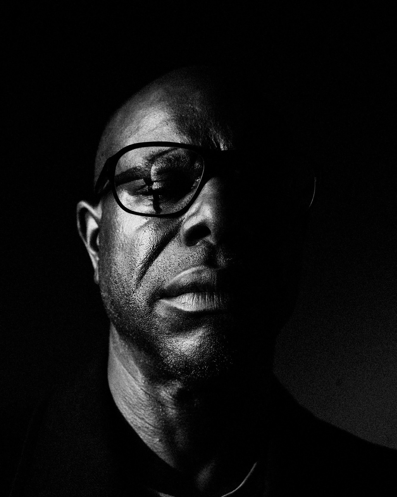 Dramatic black and white portrait of an African American man with glasses, featuring strong chiaroscuro lighting and a focused gaze.