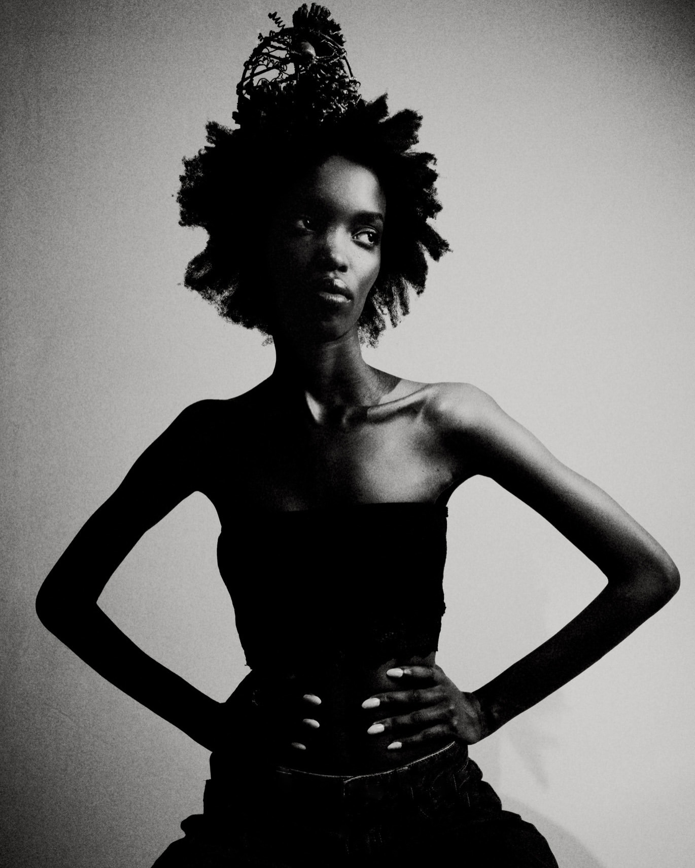 Black and white portrait of a Black woman with hands on hips, wearing a dark strapless top and decorative headpiece in a studio.