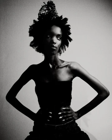Black and white portrait of a Black woman with hands on hips, wearing a dark strapless top and decorative headpiece in a studio.