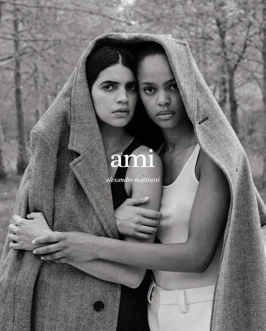 Two models in a studio shot wearing a herringbone coat over their shoulders, with a blurred forest background and the AMI logo visible.