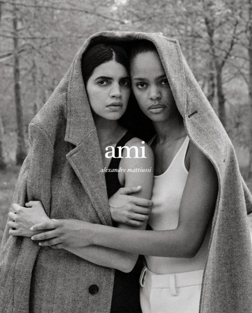 Two models in a studio shot wearing a herringbone coat over their shoulders, with a blurred forest background and the AMI logo visible.