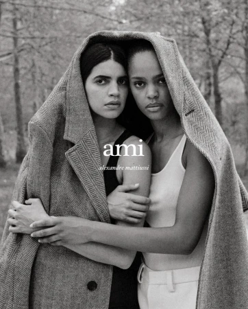 Two models in a studio shot wearing a herringbone coat over their shoulders, with a blurred forest background and the AMI logo visible.