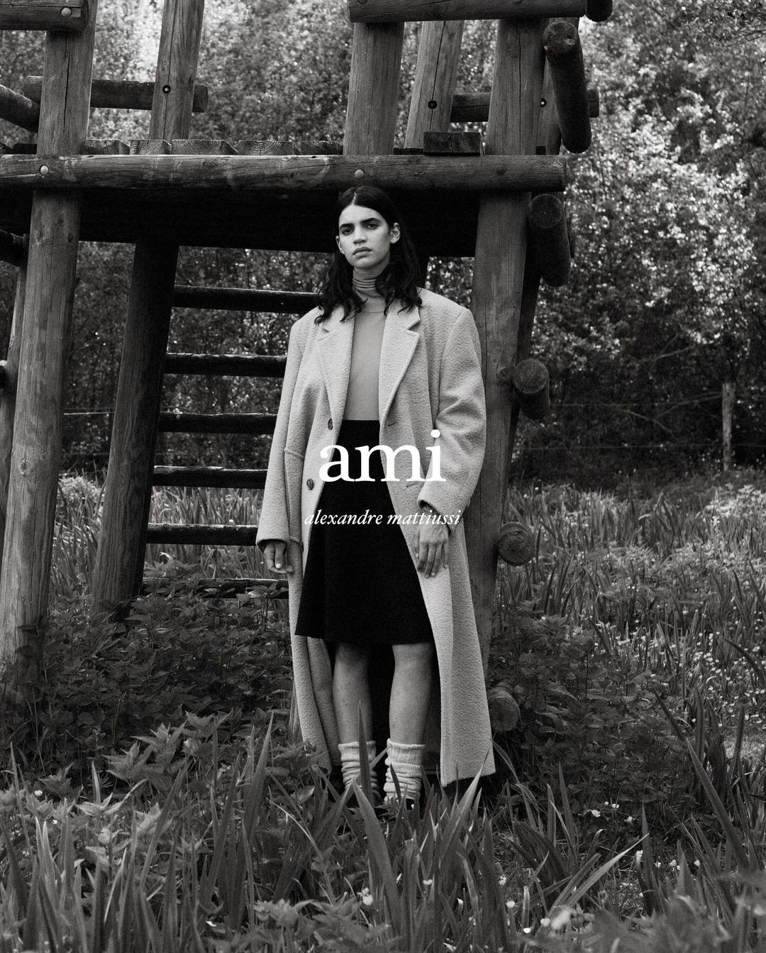 An adult female stands outside a rustic wooden tower, wearing an oversized coat, black skirt, turtleneck, and socks in black and white.