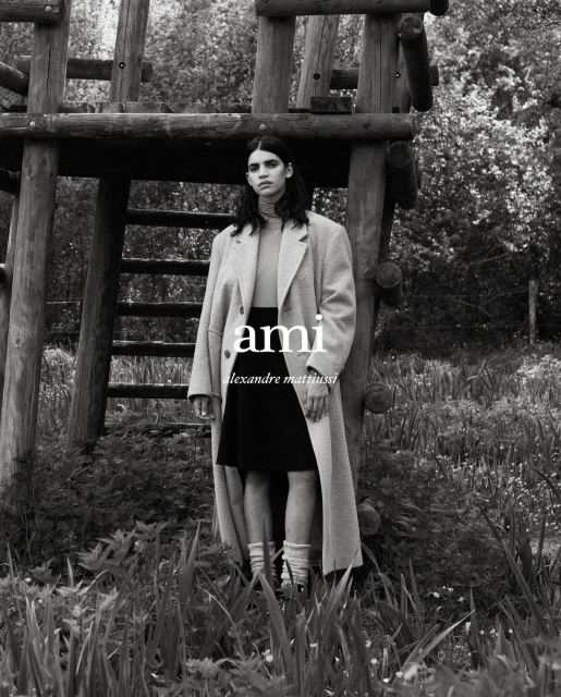 An adult female stands outside a rustic wooden tower, wearing an oversized coat, black skirt, turtleneck, and socks in black and white.